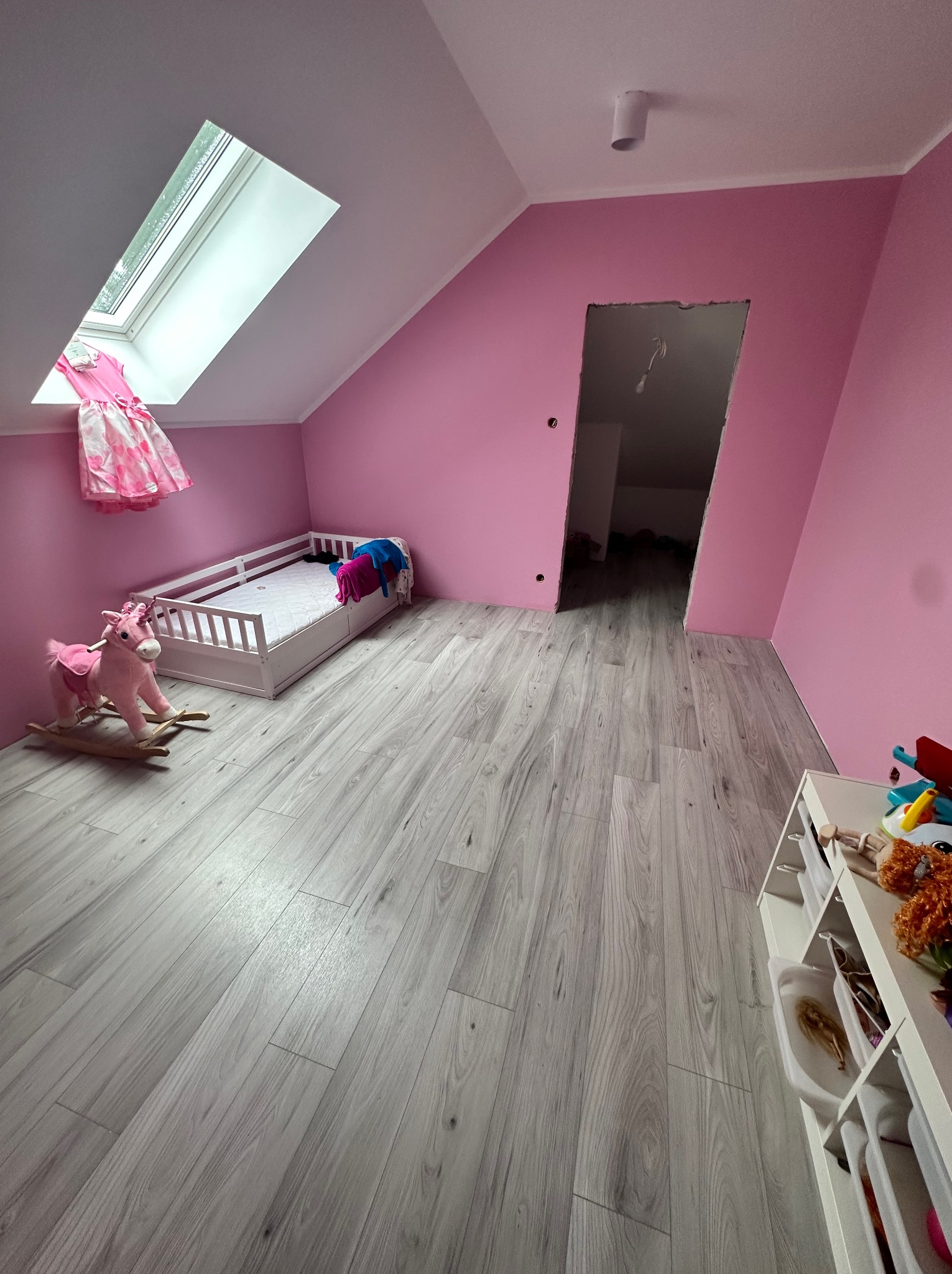 Pomalowany na różowo pokój dziecięcy na poddaszu z szarymi panelami podłogowymi, łóżkiem, konikiem na biegunach i oknem dachowym z wiszącą sukienką.