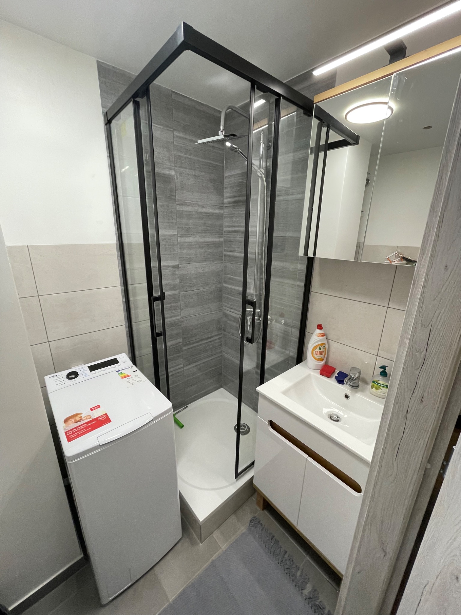 Nowoczesna łazienka z kabiną prysznicową typu walk-in z czarnymi profilami, białą umywalką z szafką, pralką i szarymi płytkami imitującymi drewno na ścianach.