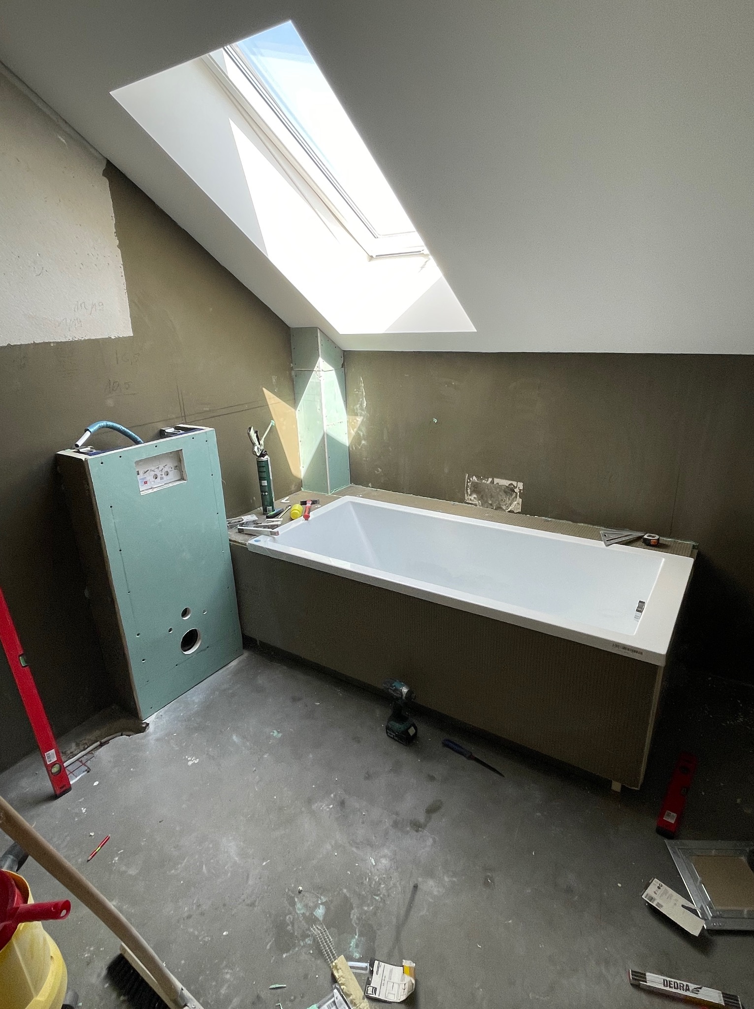 Łazienka w trakcie prac: biała wanna obudowana płytkami, zielona zabudowa podtynkowego stelaża WC, surowe ściany przygotowane do wykończenia, okno dachowe w skosie, narzędzia i akcesoria budowlane...