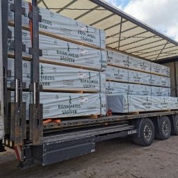 WOODPECKER PAULINA PŁOŃCZYK - Ciężarówka z odkrytą naczepą załadowana paletami z pakietami tarcicy budowlanej, zabezpieczonej folią z nadrukowanym logo producenta.