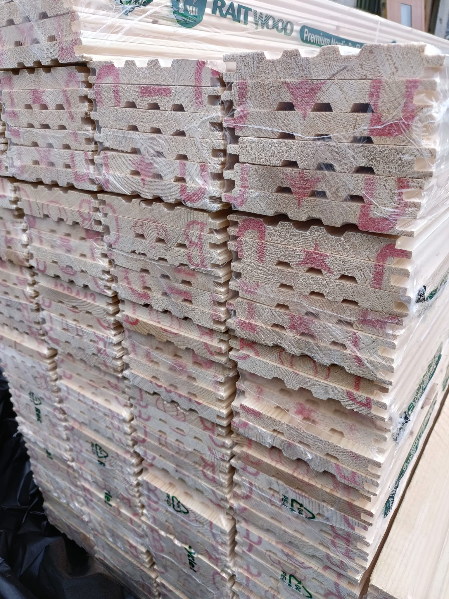 Stos ryflowanych desek boazeryjnych z jasnego drewna, zabezpieczonych folią, z widocznymi oznaczeniami producenta i recyklingu.