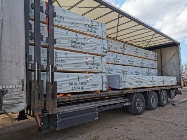 Ciężarówka z odkrytą naczepą załadowana paletami z pakietami tarcicy budowlanej, zabezpieczonej folią z nadrukowanym logo producenta.