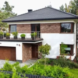 Dwupiętrowy dom z garażem, białymi ścianami i ceglaną elewacją, balkon z roślinami w donicach, zadbany ogród z drzewami i krzewami.
