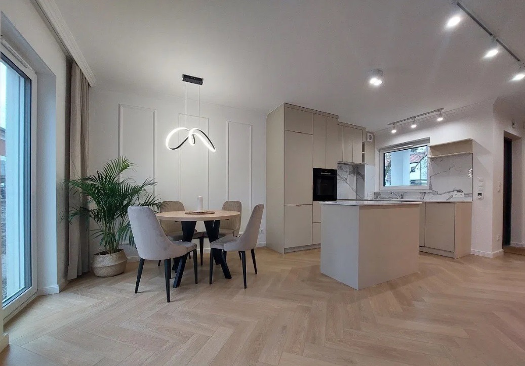Jasne wnętrze: parkiet w jodełkę, okrągły stół z krzesłami, minimalistyczna kuchnia z wyspą i nowoczesna lampa wisząca.