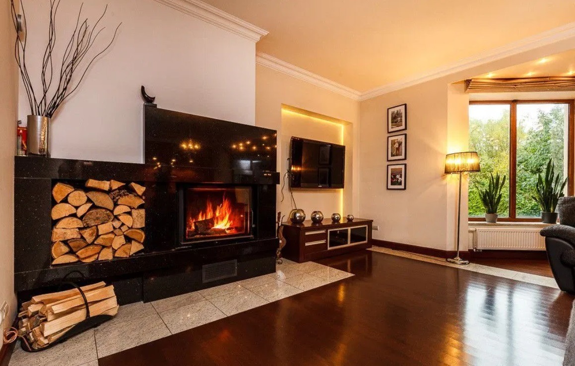 Elegancki salon z czarnym kominkiem z paleniskiem i drewnem, parkiet z ciemnego drewna, telewizor na ścianie, lampa podłogowa i rośliny doniczkowe przy oknie.