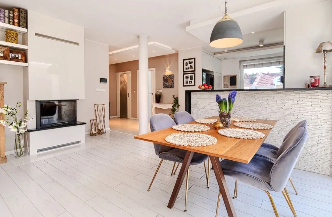 Jasne wnętrze jadalni z drewnianym stołem, aksamitnymi krzesłami na złotych nóżkach, białymi deskami podłogowymi i nowoczesnym kominkiem w tle.