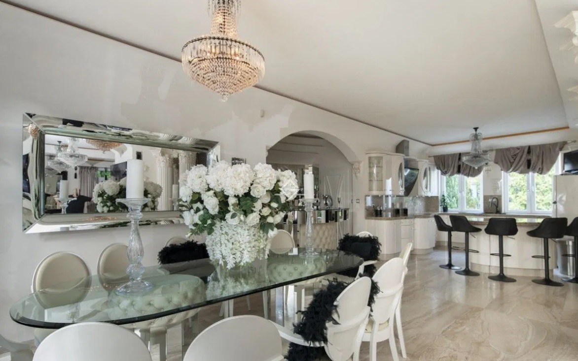 Luksusowa jadalnia w Bielsku-Białej z szklanym stołem, białymi krzesłami ozdobionymi czarnymi piórami, kryształowym żyrandolem i lustrem odbijającym wnętrze, widok na otwartą kuchnię.