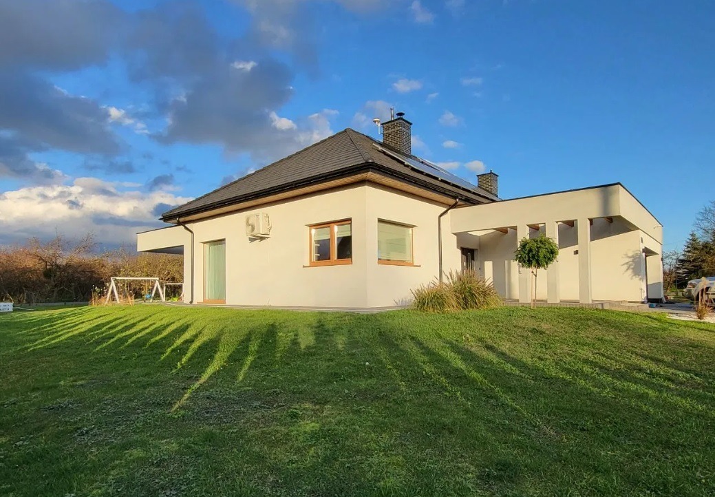 Nowoczesny, parterowy dom z białymi ścianami, ciemną dachówką i panelami słonecznymi, widziany z poziomu trawnika z wyraźnymi cieniami drzew.