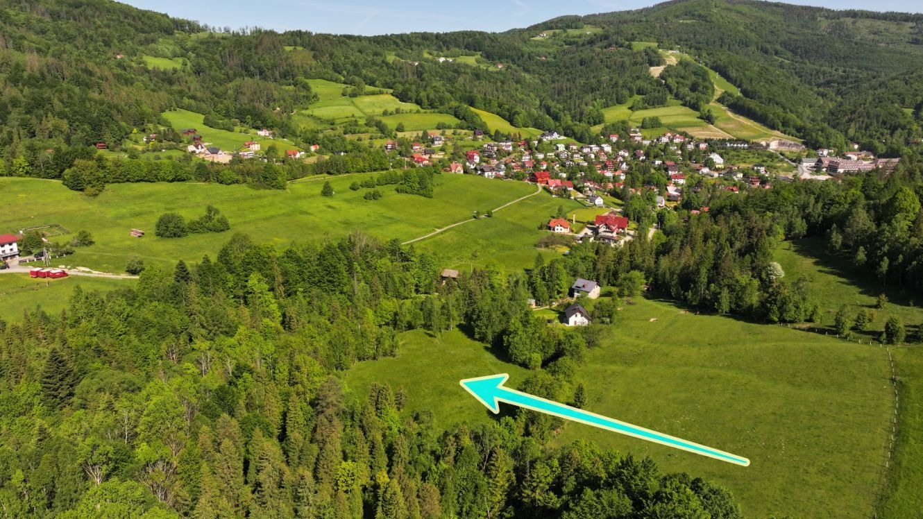 Panoramiczne ujęcie działki budowlanej na zboczu wzgórza z widokiem na wieś, zaznaczonej błękitną strzałką.