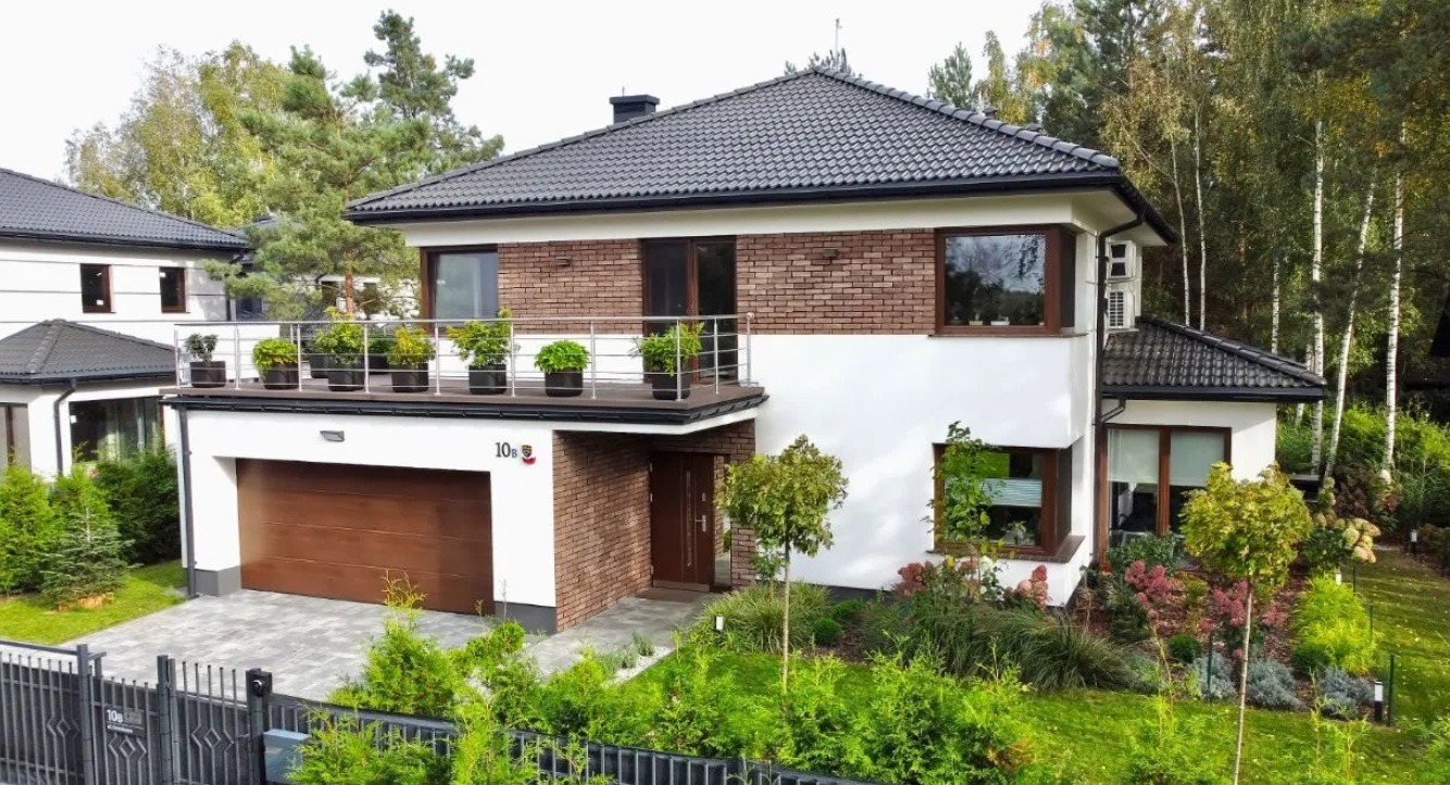Dwupiętrowy dom z garażem, białymi ścianami i ceglaną elewacją, balkon z roślinami w donicach, zadbany ogród z drzewami i krzewami.