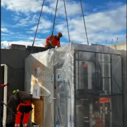 Montaż prefabrykowanej łazienki na placu budowy. Robotnicy w odblaskowych ubraniach nadzorują umieszczenie modułu za pomocą łańcuchów dźwigu.