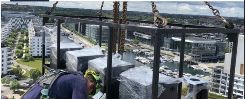 Montaż elementów wentylacyjnych na wysokości z widokiem na miasto i port jachtowy. Praca zabezpieczona uprzężą, elementy w folii ochronnej.