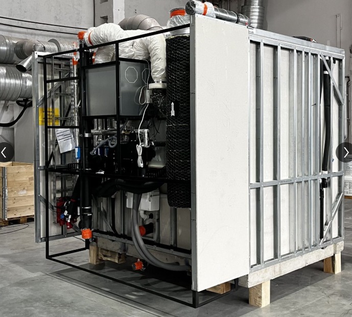 Skomplikowana instalacja techniczna w metalowej ramie, z widocznymi rurami, przewodami i elementami konstrukcyjnymi, umieszczona na drewnianych paletach w hali.