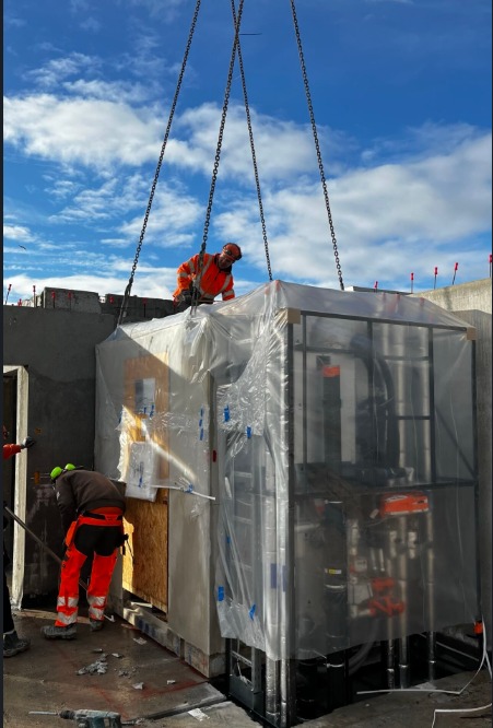 Montaż prefabrykowanej łazienki na placu budowy. Robotnicy w odblaskowych ubraniach nadzorują umieszczenie modułu za pomocą łańcuchów dźwigu.