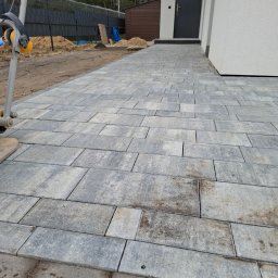 BrukStan - Ułożona szara kostka brukowa prowadząca do wejścia domu, widoczne niedociągnięcia na obrzeżach i pozostawione narzędzia na obrzeżach.