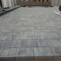 Ułożona szara kostka brukowa na posesji, widoczny fragment domu z klimatyzatorem, drabina, taczka i narzędzia brukarskie w tle.