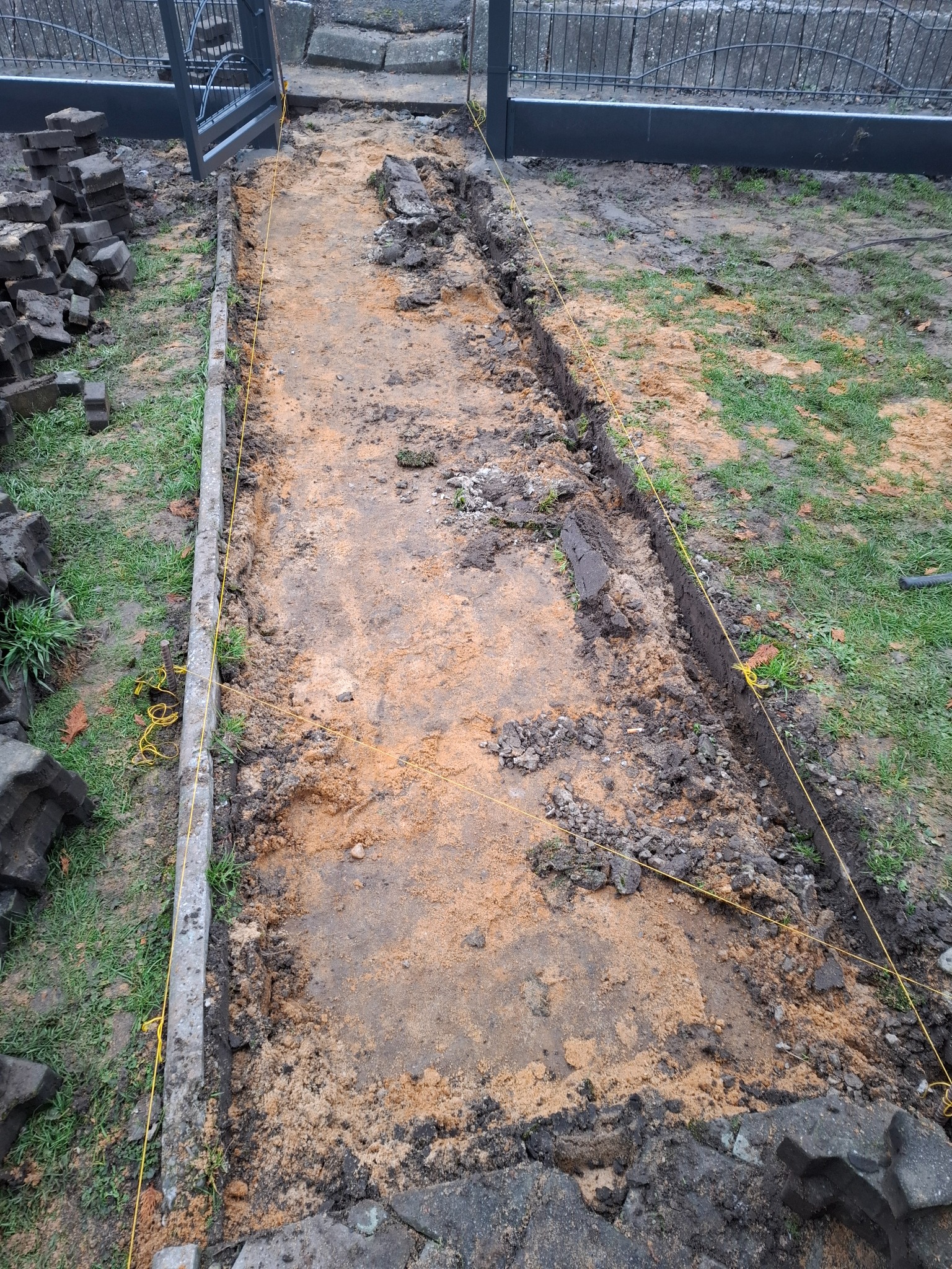 Przygotowanie podłoża pod ścieżkę z kostki brukowej: wytyczony pas ziemi z piaskiem, obrzeża betonowe, stos kostki brukowej obok, widoczne sznurki wyznaczające granice.