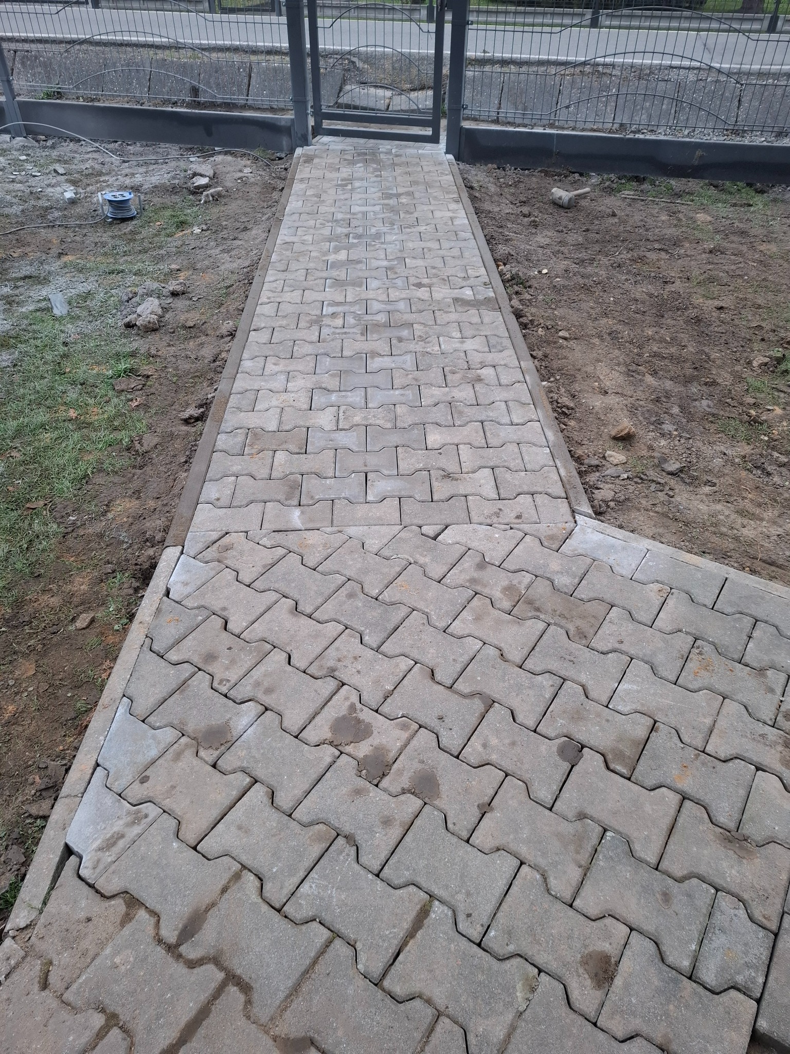 Świeżo ułożony chodnik z szarej kostki brukowej typu 'I', z widocznymi niedociągnięciami przy łączeniu z istniejącą ścieżką i nierównościami terenu po bokach, prowadzący do metalowej bramki.