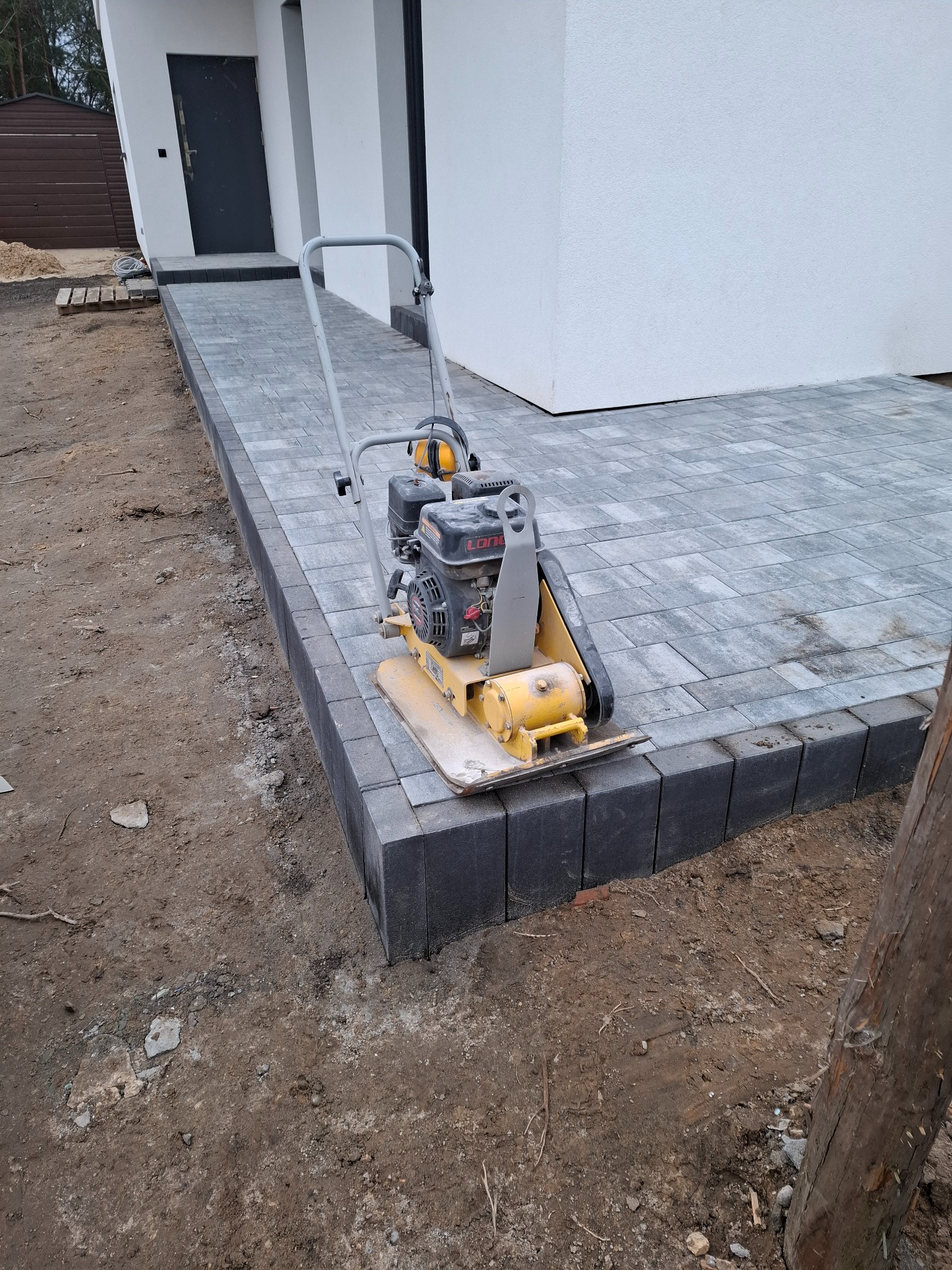 Żółta zagęszczarka spalinowa stoi na nowo ułożonej szarej kostce brukowej przed budynkiem z białym tynkiem. Widoczne ślady ziemi na obrzeżach kostki.