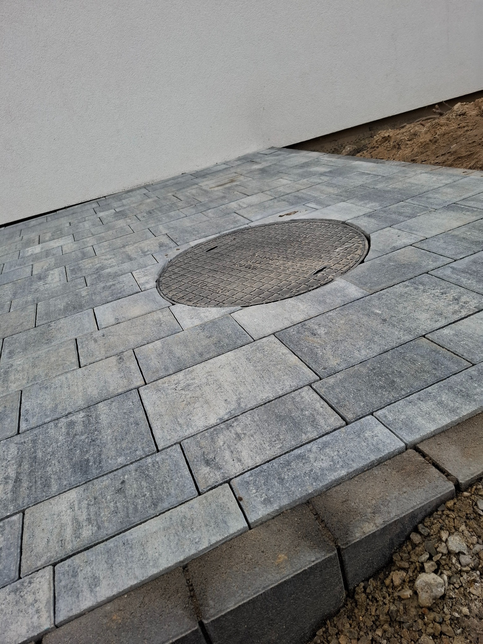 Szara kostka brukowa ułożona przy białej ścianie budynku, z okrągłą studzienką kanalizacyjną wkomponowaną w nawierzchnię, widoczne obrzeża i podsypka.