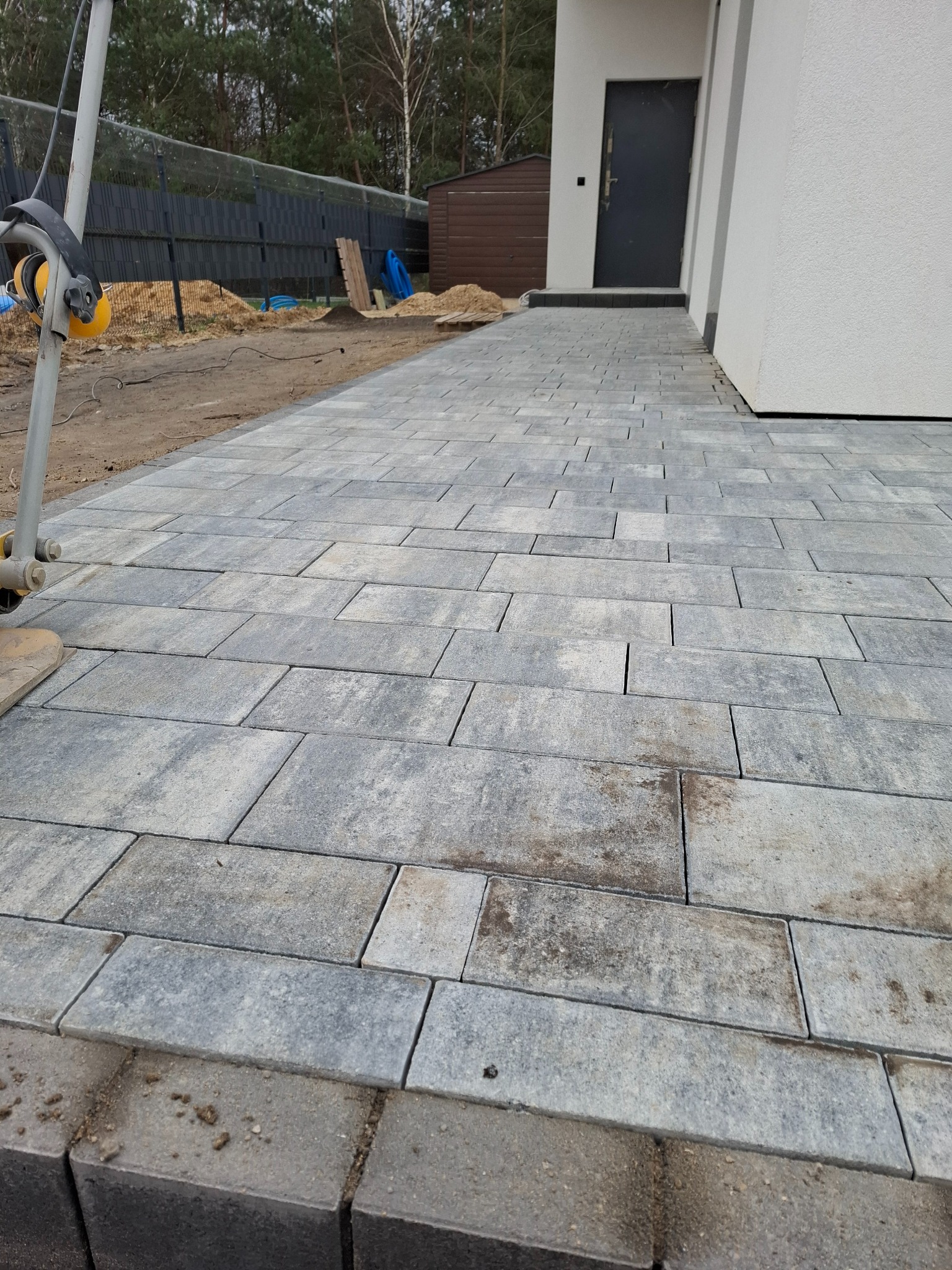 Ułożona szara kostka brukowa prowadząca do wejścia domu, widoczne niedociągnięcia na obrzeżach i pozostawione narzędzia na obrzeżach.