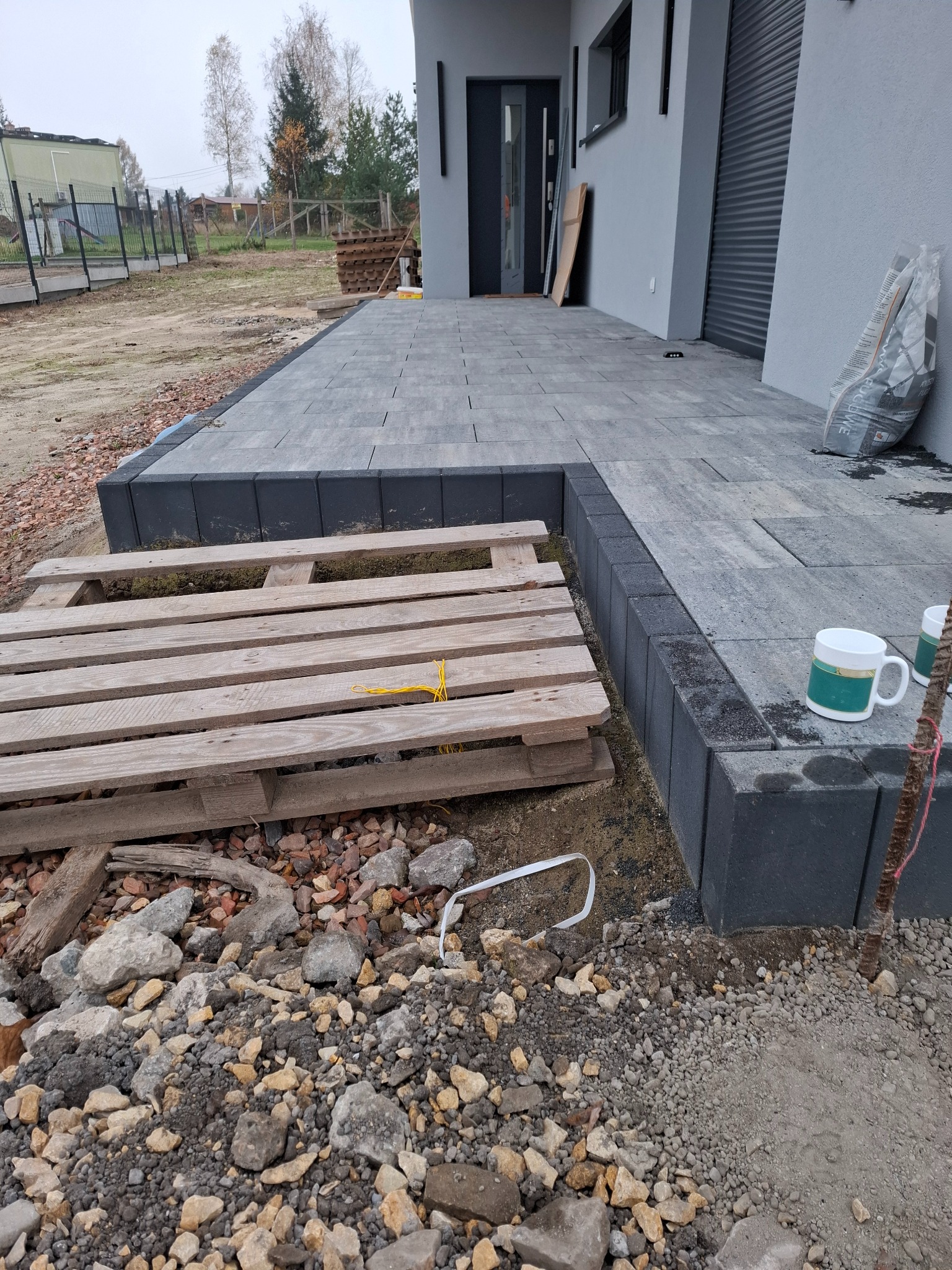 Układanie szarej kostki brukowej w Gliwicach, widoczna paleta z materiałami budowlanymi i kubki na krawędzi.