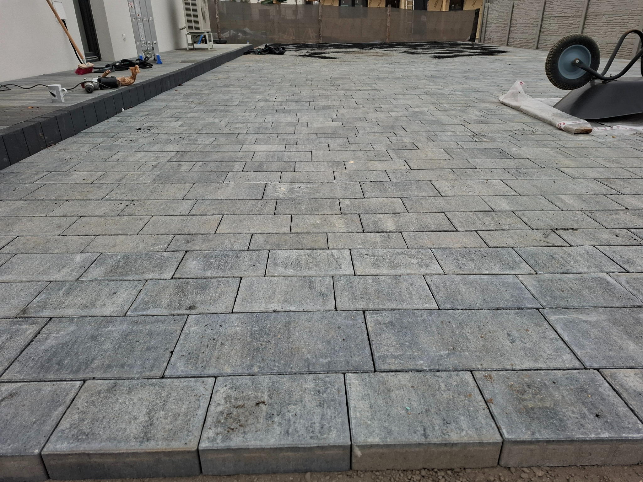 Ułożona szara kostka brukowa na posesji, widoczny fragment domu z klimatyzatorem, drabina, taczka i narzędzia brukarskie w tle.