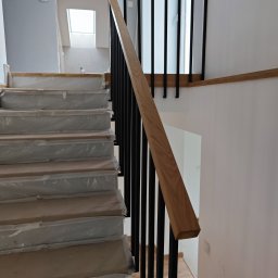 Mateusz Stec - Nowoczesne schody z drewnianymi stopniami zabezpieczonymi folią i czarną metalową balustradą z drewnianą poręczą, widok z dołu.