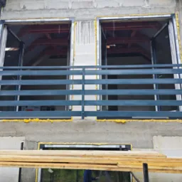 Szara balustrada balkonowa z poziomymi szczeblami, zamontowana na surowym balkonie z oknami. Widoczne pianki montażowe i drewniane deski rusztowania.