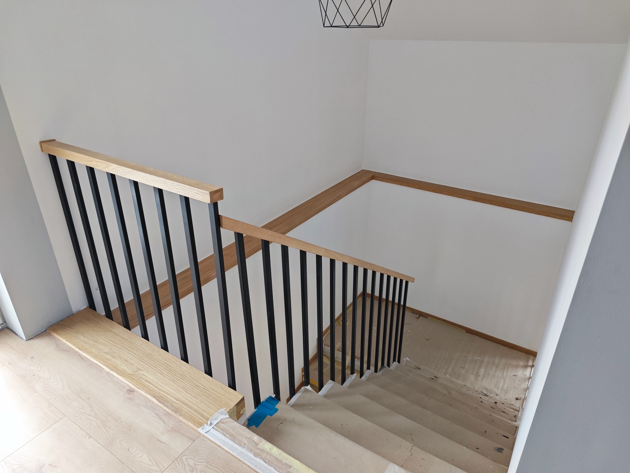 Nowoczesne schody z drewnianymi stopniami i czarną metalową balustradą, widok z góry, zabezpieczone folią malarską podczas prac wykończeniowych.