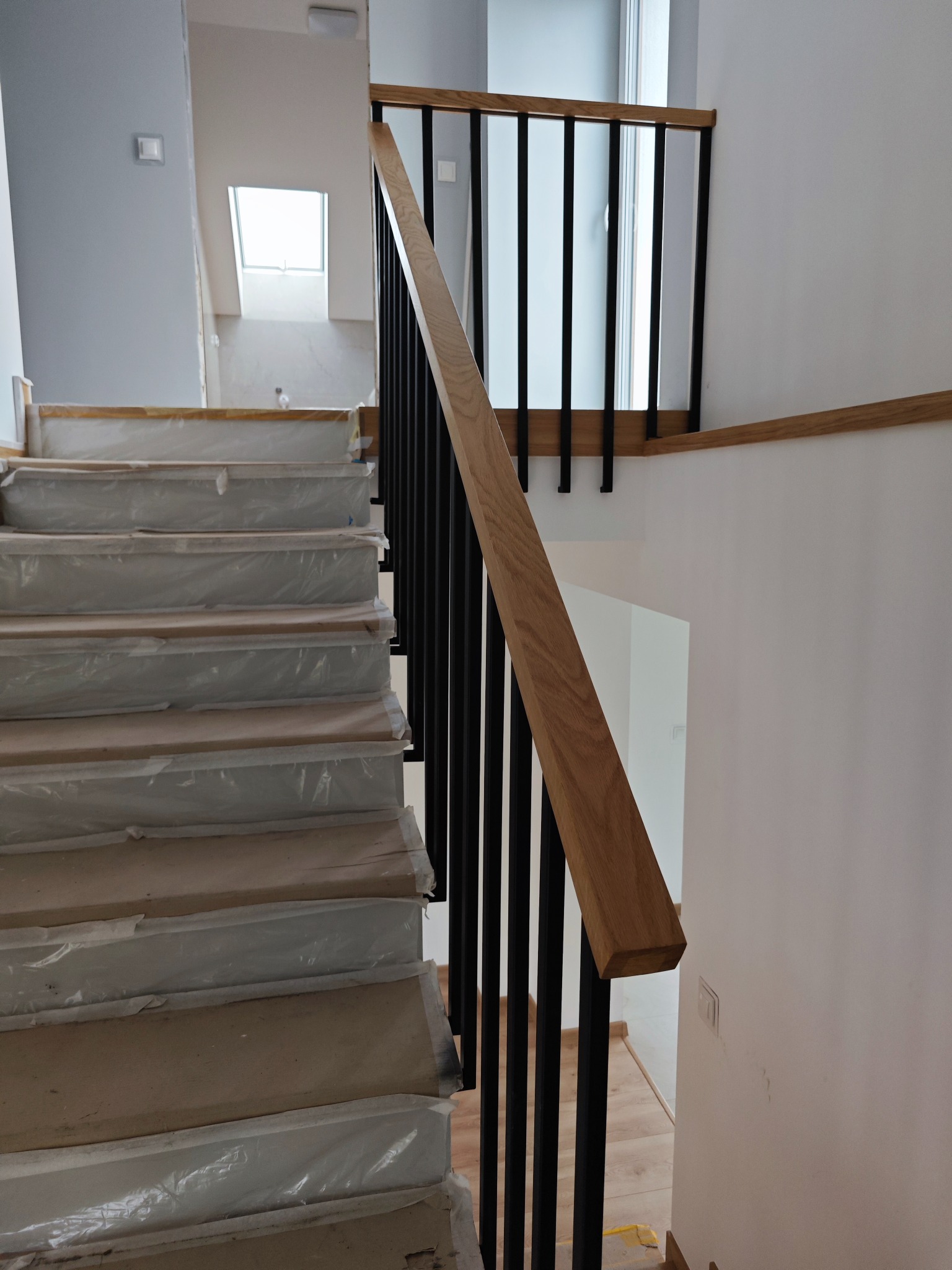 Nowoczesne schody z drewnianymi stopniami zabezpieczonymi folią i czarną metalową balustradą z drewnianą poręczą, widok z dołu.