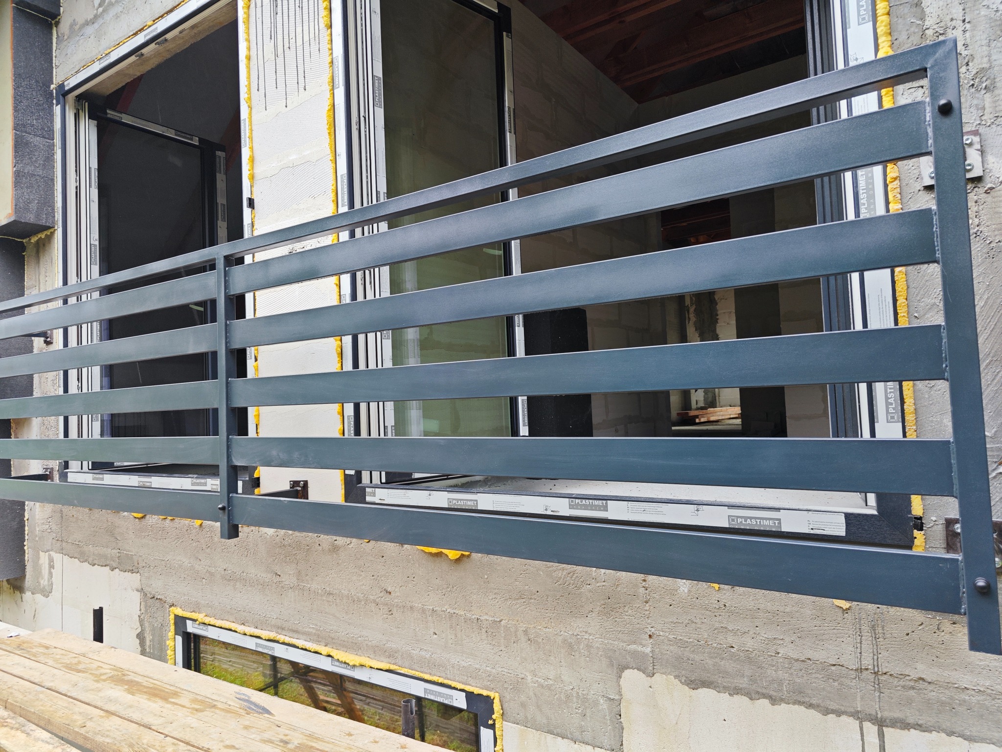 Nowoczesna, pozioma, metalowa konstrukcja zabezpieczająca balkon w surowym stanie, z widocznymi oknami i elementami konstrukcyjnymi budynku.