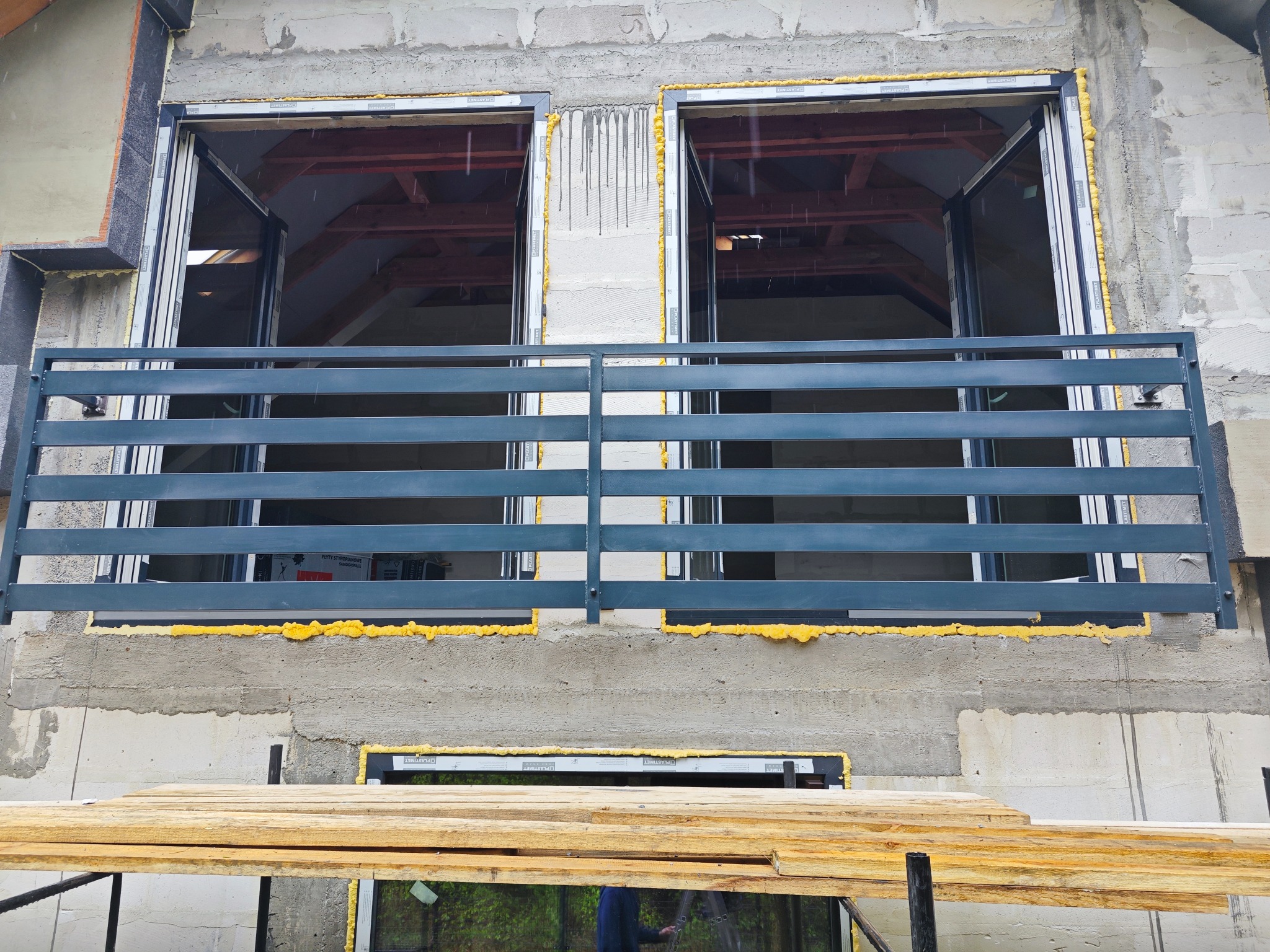 Szara balustrada balkonowa z poziomymi szczeblami, zamontowana na surowym balkonie z oknami. Widoczne pianki montażowe i drewniane deski rusztowania.