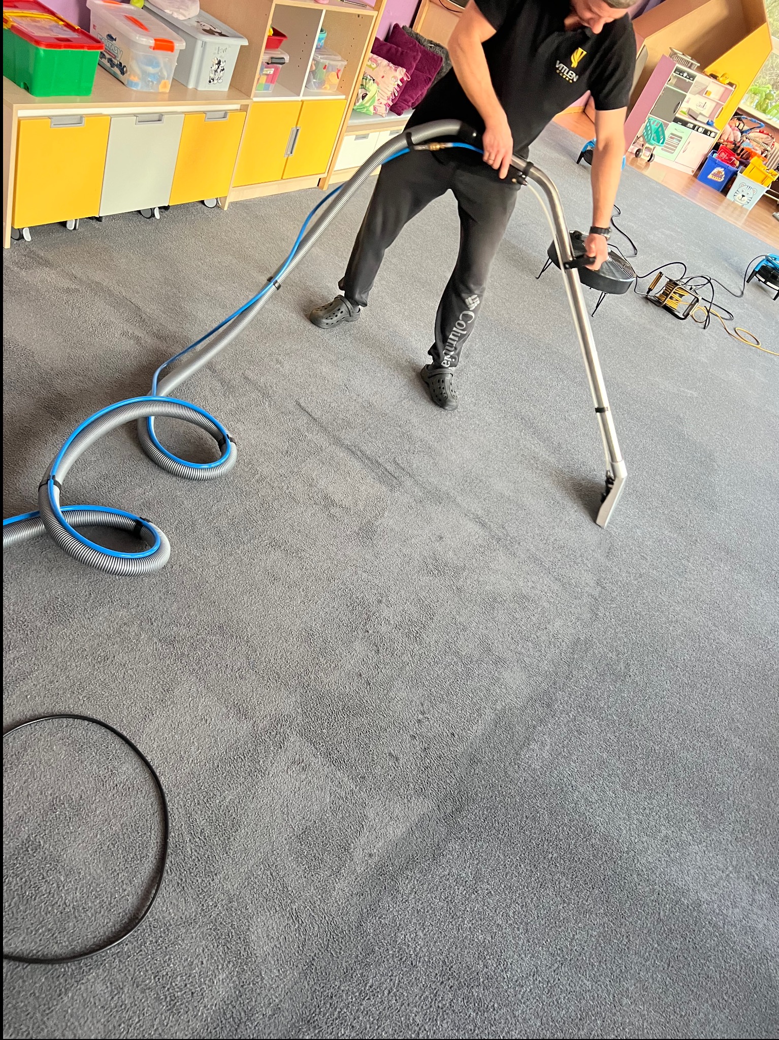 Mężczyzna czyści szary dywan w pokoju dziecięcym za pomocą profesjonalnego sprzętu. Widoczne zabawki i szafki w tle. Charakterystyczny wąż odkurzacza.