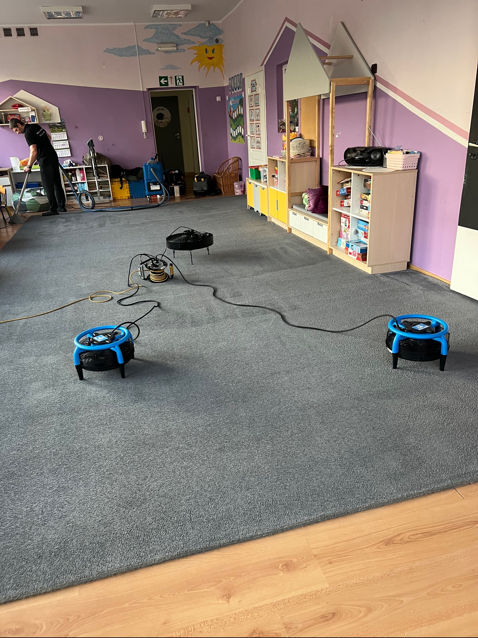 Szary dywan w pokoju zabaw przedszkola, suszony przez wentylatory po praniu. W tle osoba obsługująca odkurzacz piorący i kolorowe meble.