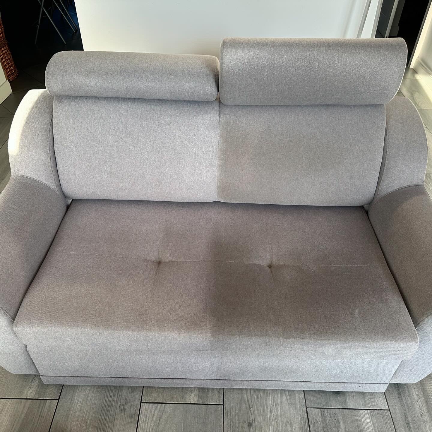 Szara kanapa po praniu tapicerki, widoczna różnica w czystości między wypraną a niewypraną częścią. Jasne tło, drewnopodobna podłoga.