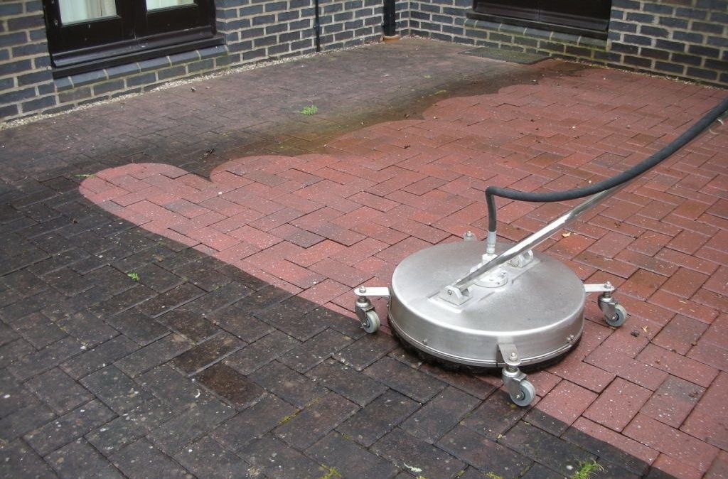 Mycie kostki brukowej: fragment wyczyszczonej czerwonej kostki brukowej w jodełkę kontrastuje z brudną, ciemną częścią. Widoczna maszyna czyszcząca z dyszą.