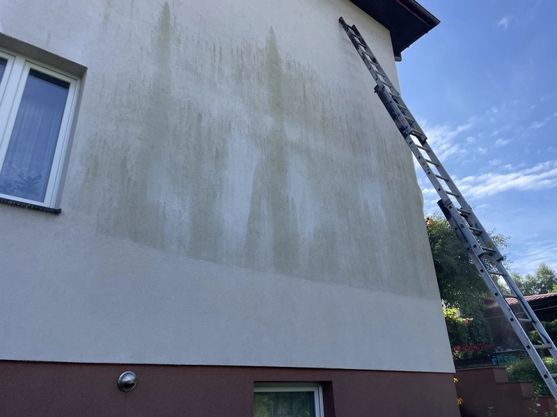 Ściana budynku z widocznymi śladami zabrudzeń i częściowo umytą powierzchnią, z drabiną opartą o ścianę. Widoczne okna i fragment dachu.