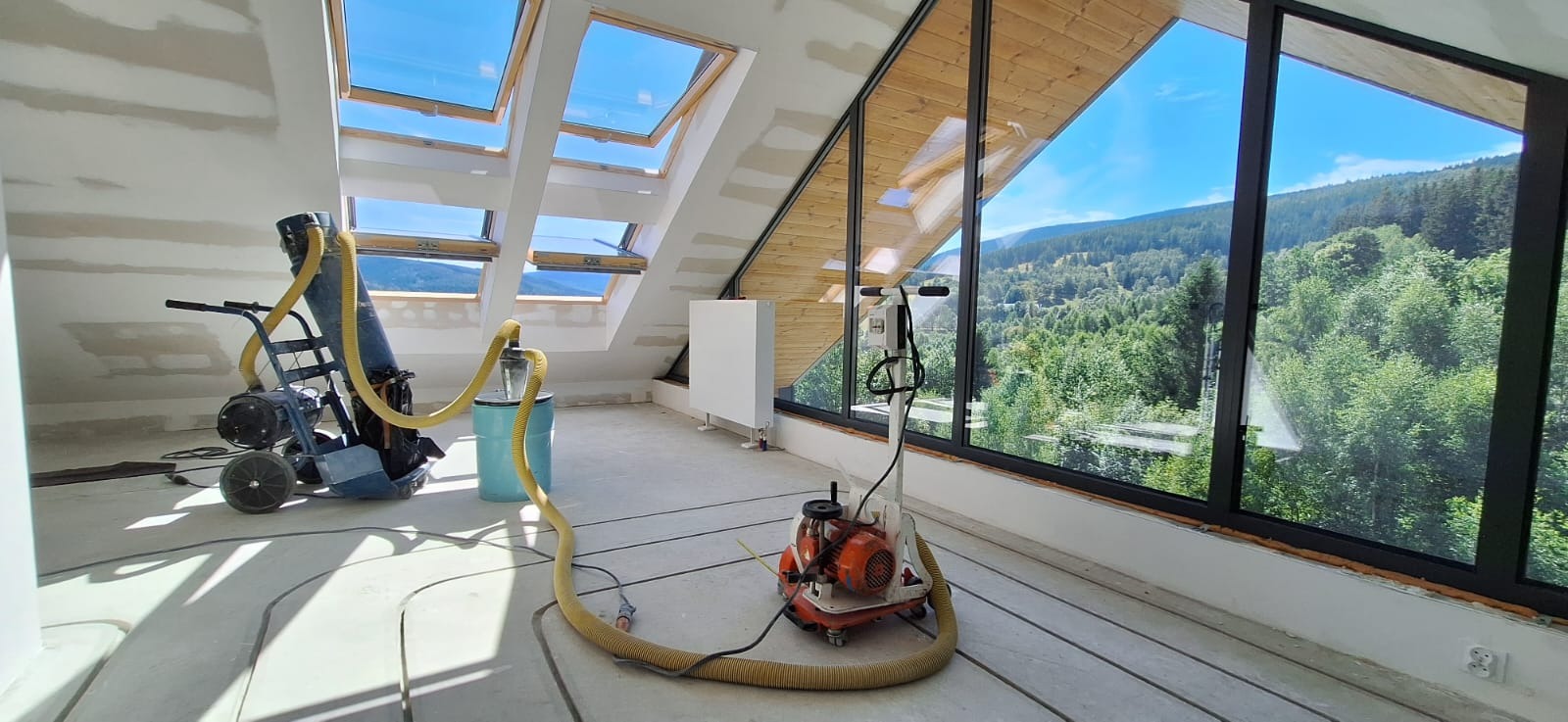 Montaż rur w systemie ogrzewania podłogowego na poddaszu z widokiem na las przez duże okno, w tle maszyny do wylewki.