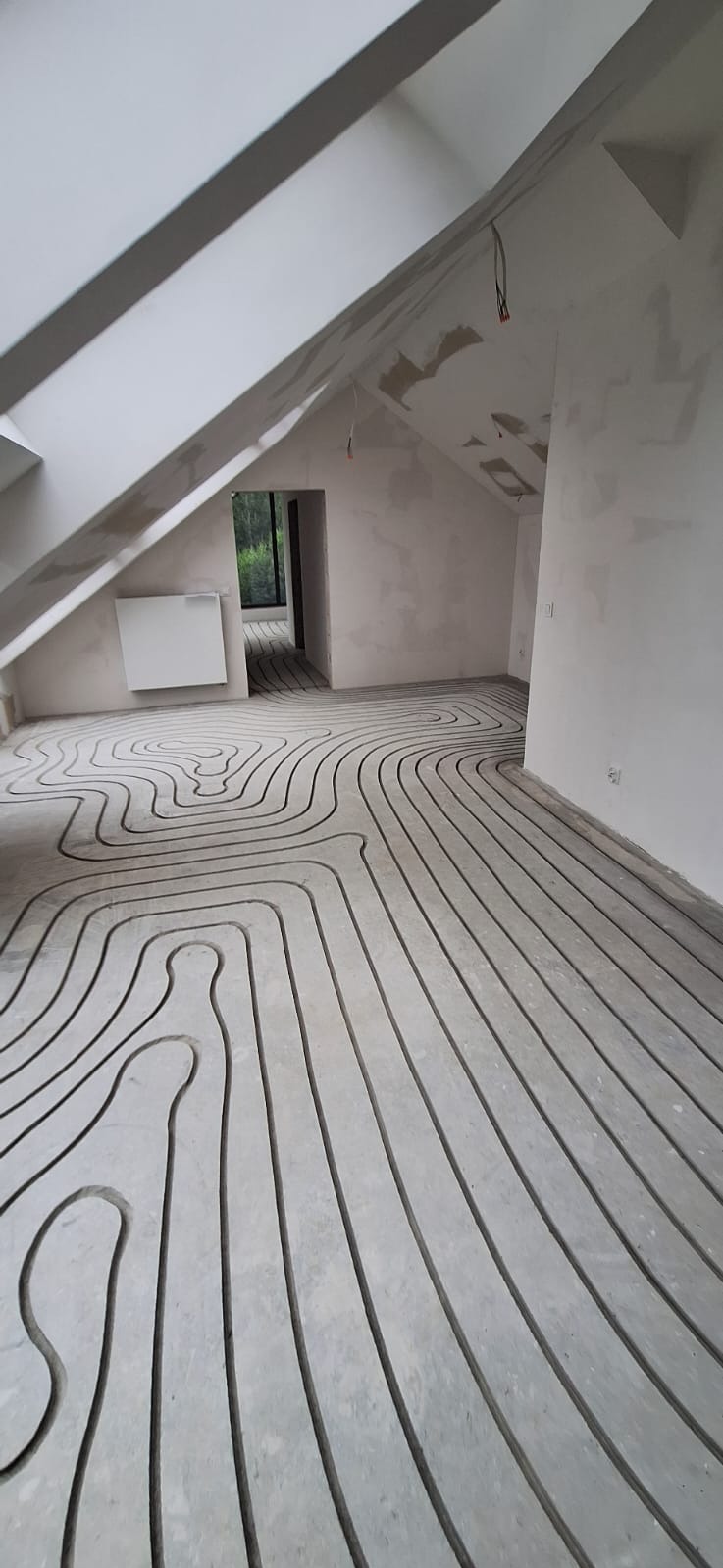 Szare rurki ogrzewania podłogowego ułożone w zygzakowaty wzór na betonowej wylewce w surowym, nieotynkowanym pomieszczeniu na poddaszu ze skosami i oknem dachowym.