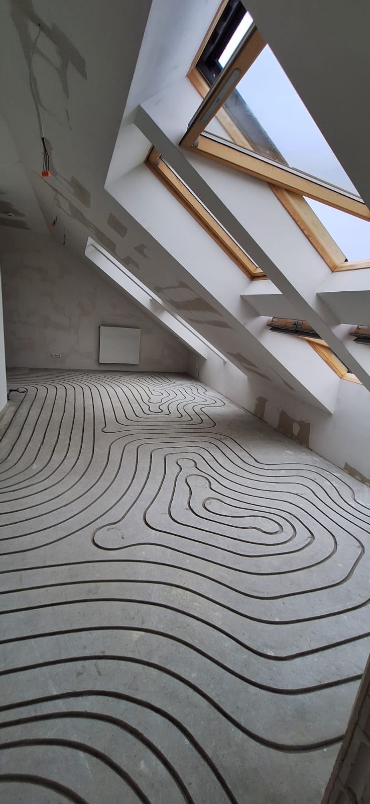 Surowy beton z wyfrezowanymi kanałami pod system ogrzewania podłogowego w jasnym pomieszczeniu ze skosami i oknami dachowymi.