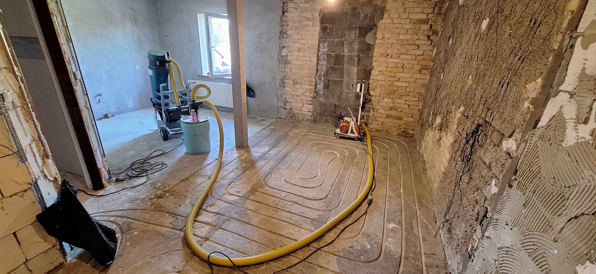 Instalacja ogrzewania podłogowego w surowym wnętrzu: widoczne rury grzewcze w wylewce, maszyna do wylewania betonu oraz fragment odsłoniętej ceglanej ściany.