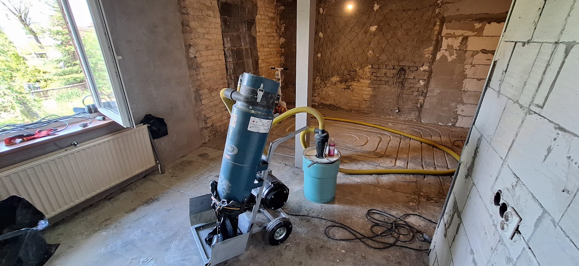 Przygotowanie do wylewki na instalacji ogrzewania podłogowego, widoczny odkurzacz przemysłowy z żółtym wężem, rury ogrzewania podłogowego rozłożone na podłodze, surowe ściany w tle.