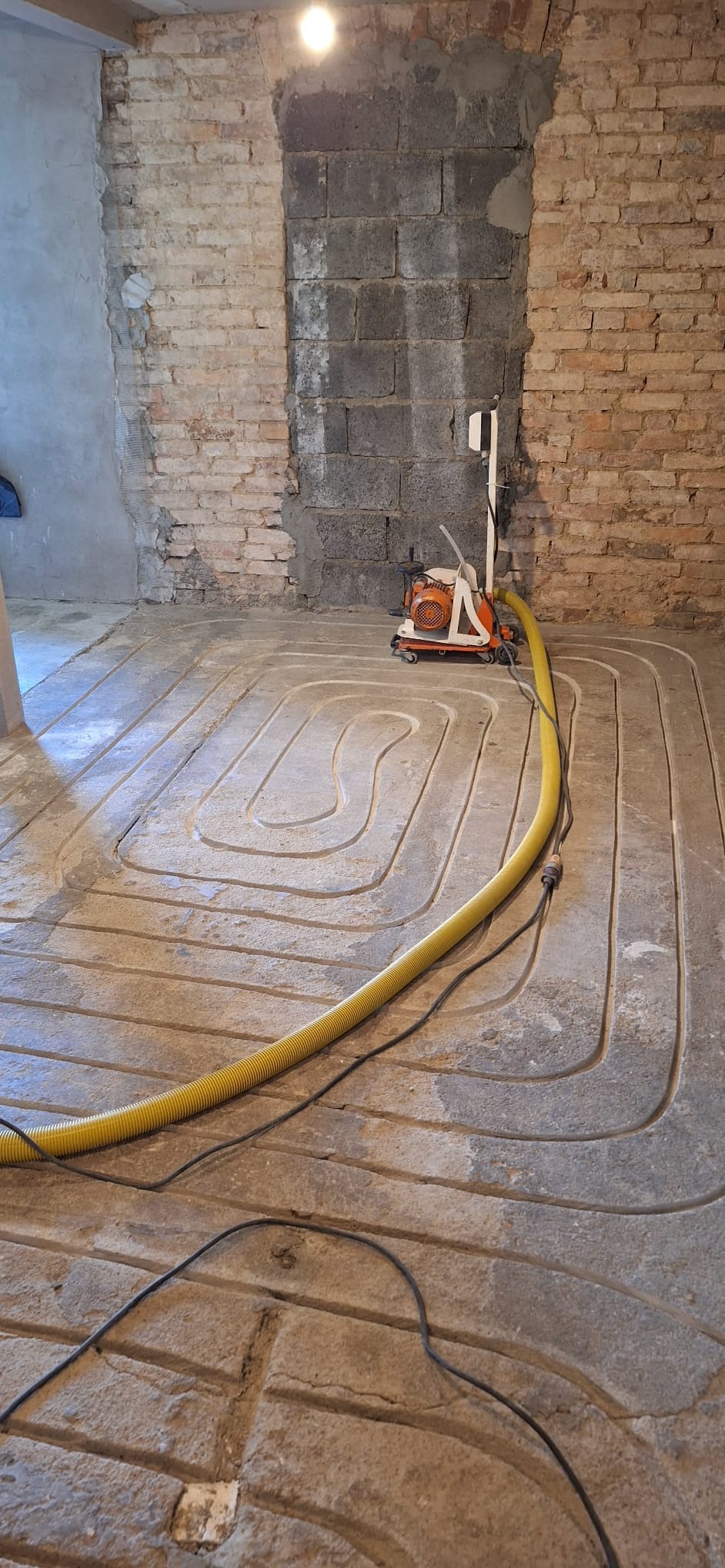 Frezowanie podłoża pod instalację ogrzewania podłogowego; widoczne wyfrezowane rowki w jastrychu, żółty wąż od odkurzacza przemysłowego i frezarka na kółkach przy ścianie z mieszanką cegieł i betonu.