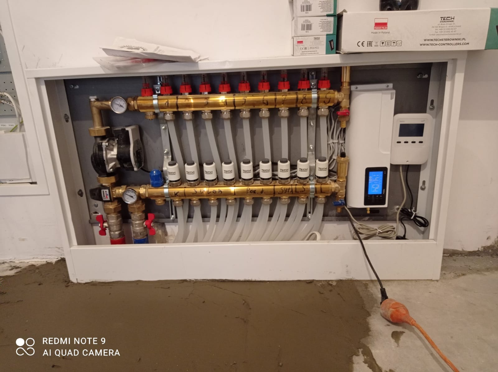 Rozdzielacz ogrzewania podłogowego w białej obudowie, widoczny na tle surowej posadzki, z przewodami i elementami sterującymi marki TECH, z widocznymi napisami 'K. SALON', 'Por. d.', 'SYPIAL.'