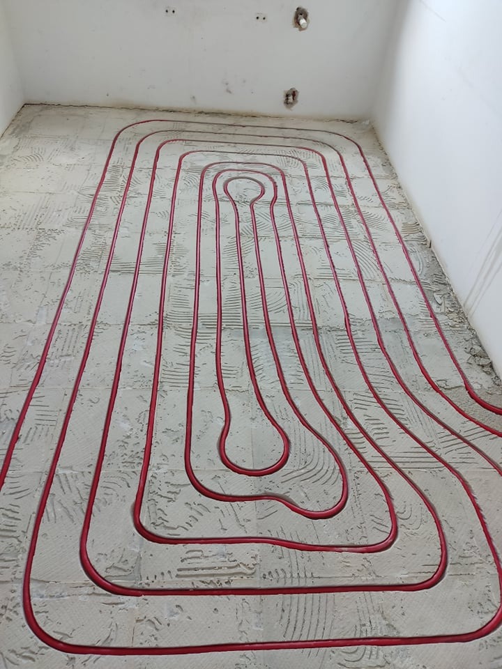 Czerwone rurki instalacji ogrzewania podłogowego ułożone na szarej, surowej posadzce w trakcie montażu, widoczne ślady po zaprawie.