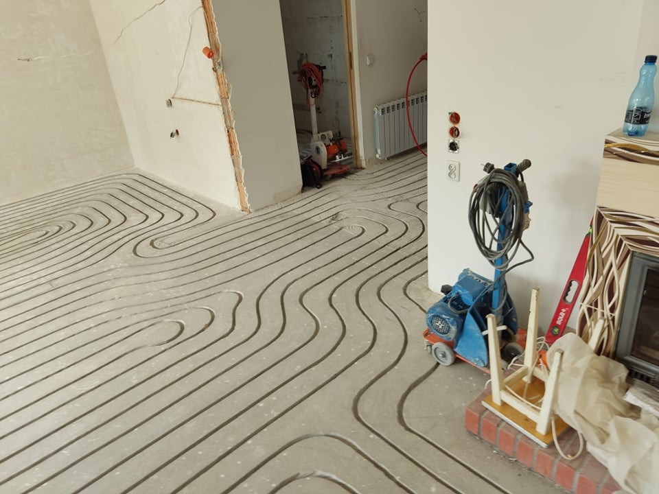 Świeżo wylany beton z zatopionymi rurami w kształcie fal, instalacja przed wyschnięciem, widoczny sprzęt do wygładzania betonu i poziomica.