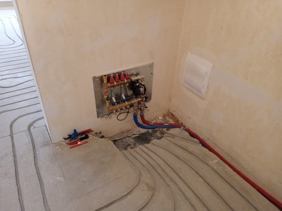 Rozdzielacz z pompą obiegową na ścianie, rury instalacji podłogowej w warstwie wylewki, narzędzia na podłodze, surowy stan wykończenia wnętrza.