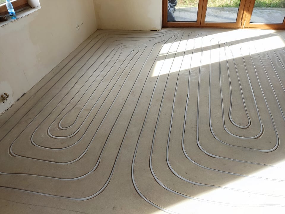 Rury układane w pętlach na podłożu, przygotowane do zalania w systemie ogrzewania, widoczne okno balkonowe w tle.