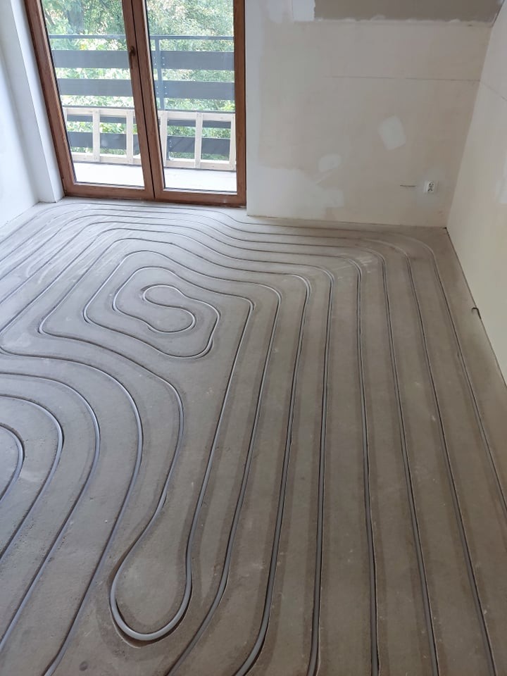 Szare rury układane w pętli na podłodze, przygotowane do zalania betonem, instalacja w trakcie montażu, widok z góry, okno balkonowe w tle.